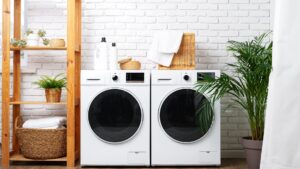 A white washer and dryer standing side by side in a bright laundry room with baskets and plants
