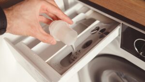 A hand pouring liquid detergent from a measuring cap into the open dispenser drawer of a washing machine