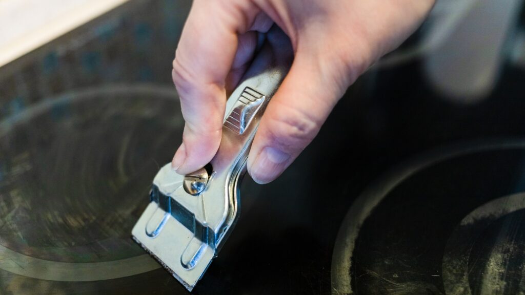 A cooktop scraper held at a shallow angle removing burnt residue from a glass surface