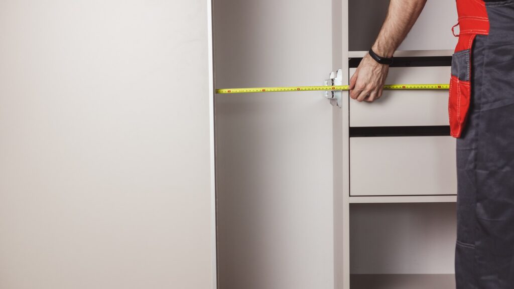 A tape measure stretched across the height of an empty kitchen cabinet opening for an oven